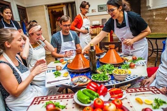 Colorful Moroccan cooking scene with tagines, spices, fresh herbs, and hands preparing food.