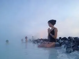 Woman sitting on rocks by a water body, with eyes closed and hands in a prayer position, during foggy weather.