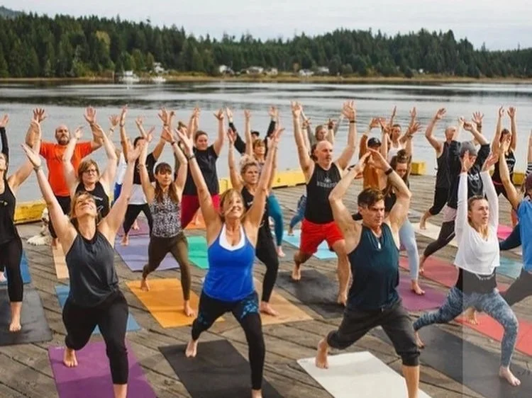 Group of people practicing yoga outdoors on a dock by a lake with trees in the background.