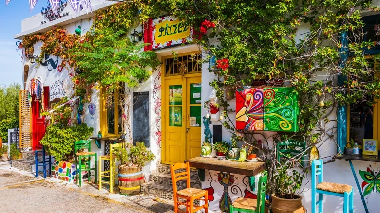 Colorful building with yellow doors, vibrant chairs, lush greenery, and decorative art, resembling a lively cafe or shop with a whimsical, artistic vibe.