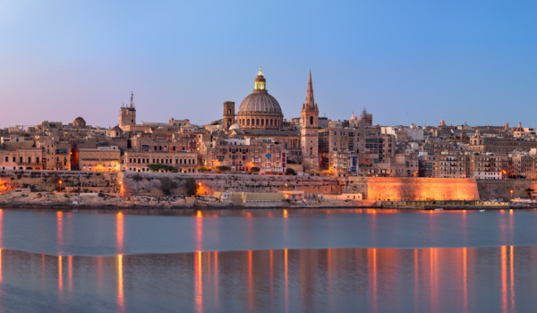 City skyline with historic buildings and domed cathedral reflecting in the water at sunset.