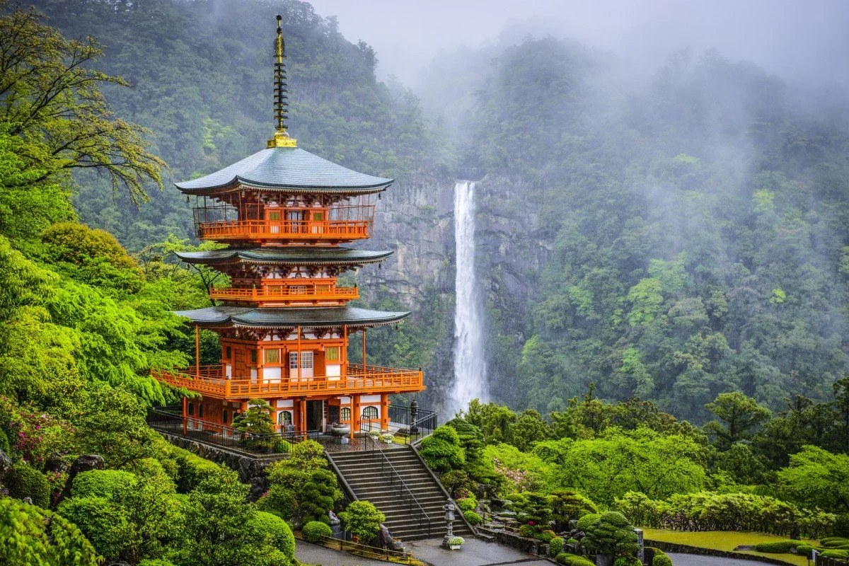 Traditional Japanese pagoda surrounded by green trees with a waterfall in the misty background