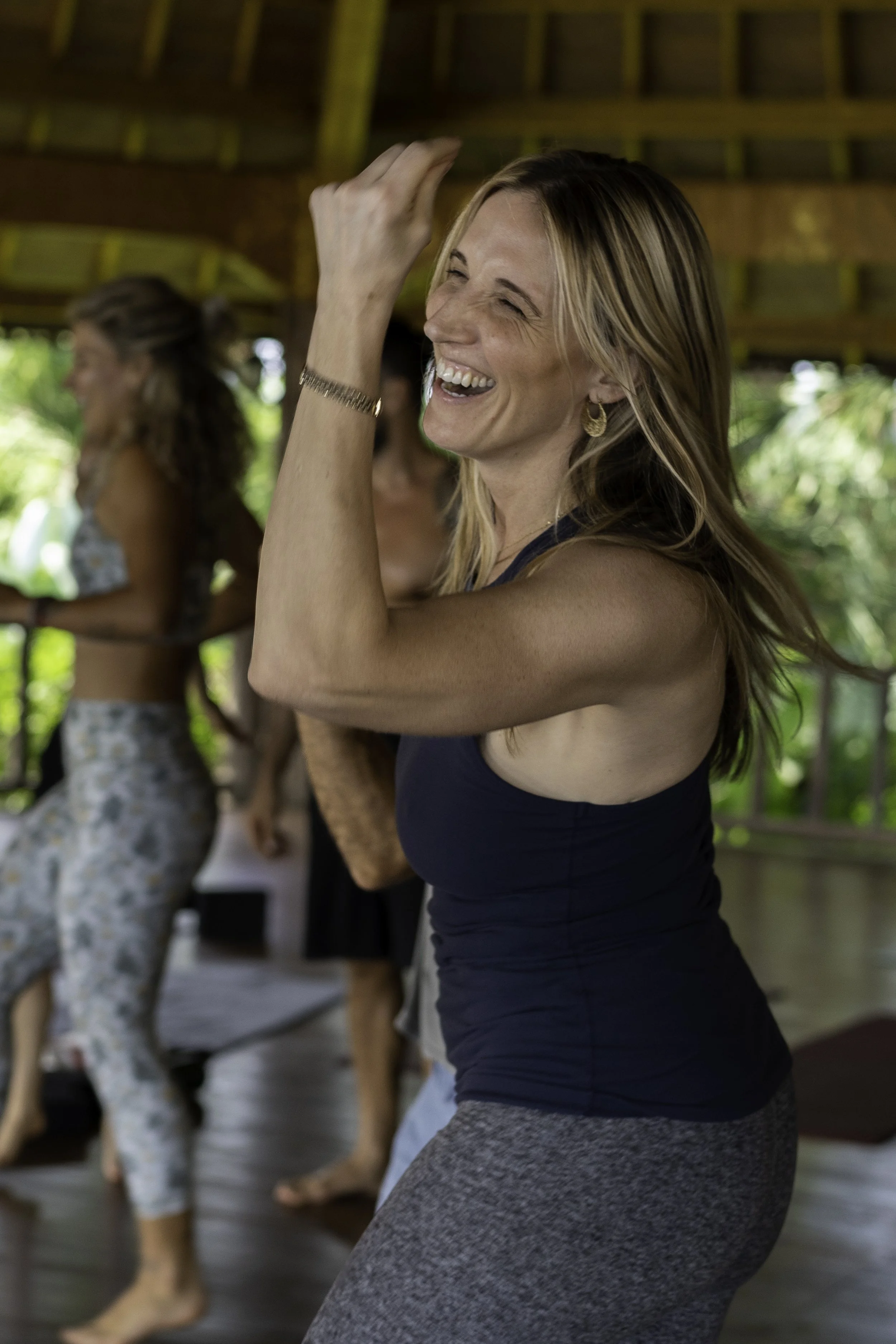 A woman with blonde hair, wearing a dark tank top and gray leggings, smiling and raising her hand in a yoga or dance class. Other women are behind her, also participating in the activity, in a wooden, open-air studio with greenery outside.