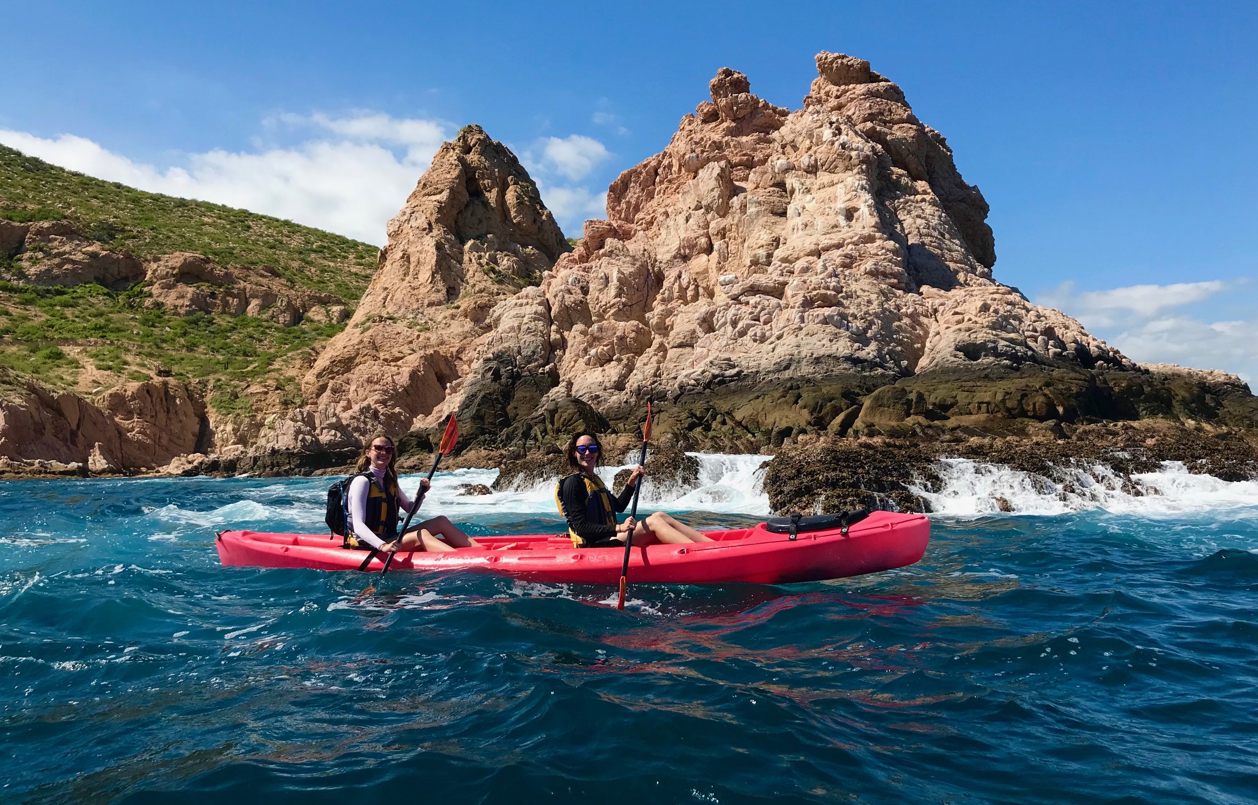 Two women in a red kayak paddling in the ocean near rocky cliffs with green hills and a partly cloudy sky.