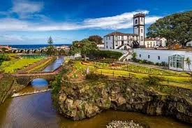 A scenic view of a historic town with a river running through it, a stone bridge, green parks, and a church with a tall clock tower under a blue sky with clouds.
