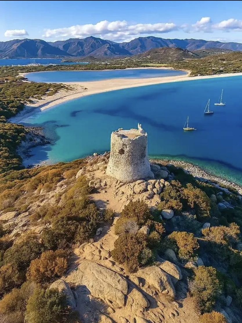 A scenic coastal landscape with a rocky hill featuring a historic stone tower, overlooking a bay with turquoise waters, sandy beaches, sailboats, lush greenery, and distant mountains under a partly cloudy sky.