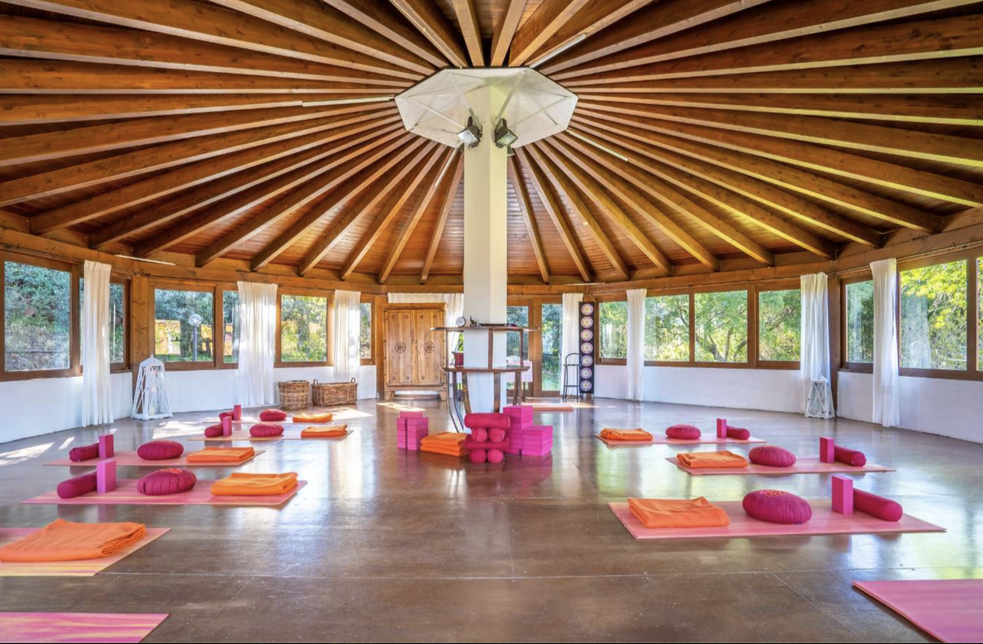 Interior of a wooden dome-shaped yoga or meditation studio with windows, pink yoga mats, orange towels, round pink cushions, and pink blocks arranged for practice or meditation.