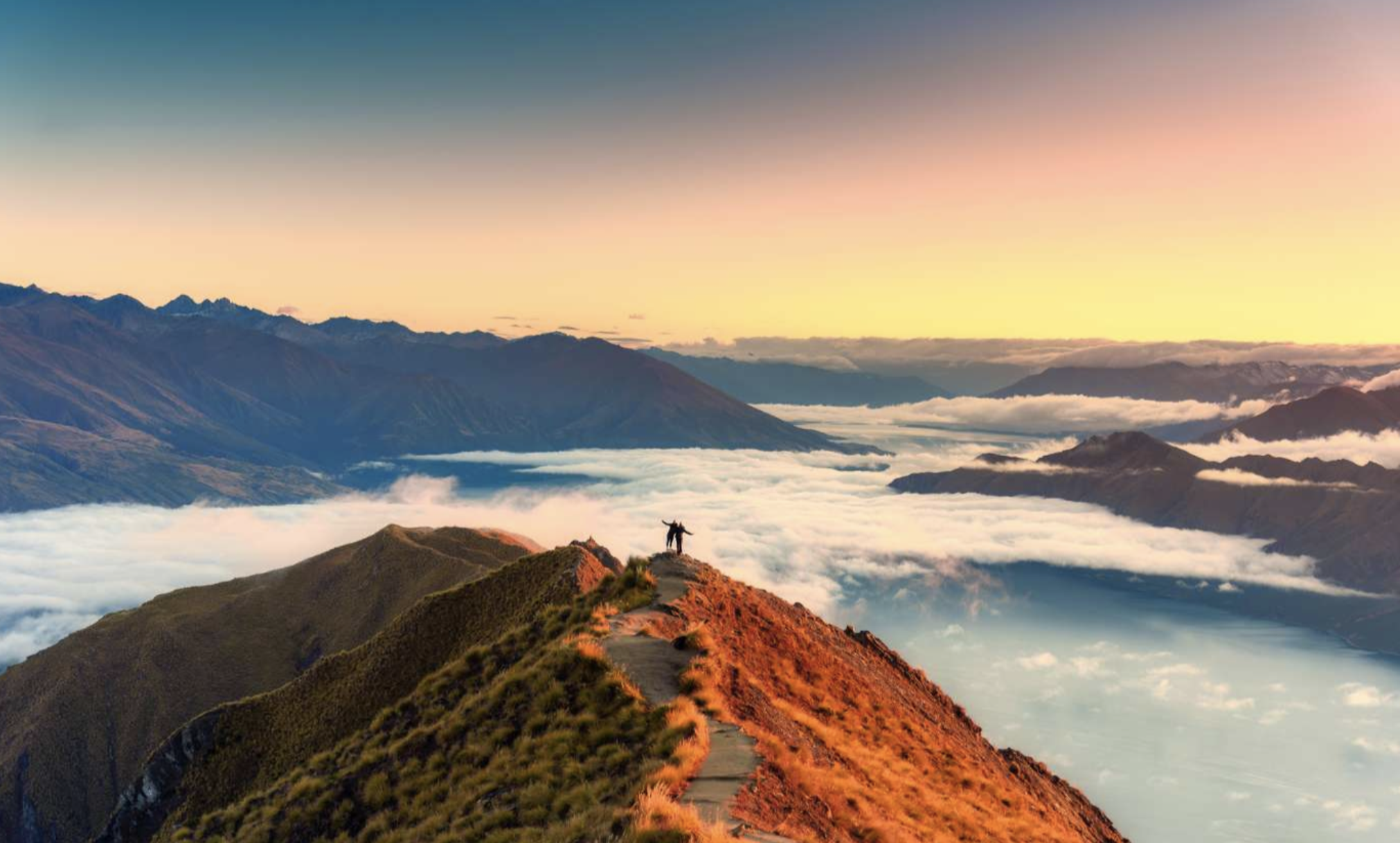 A person standing on a mountain peak overlooking a valley filled with clouds at sunrise or sunset.