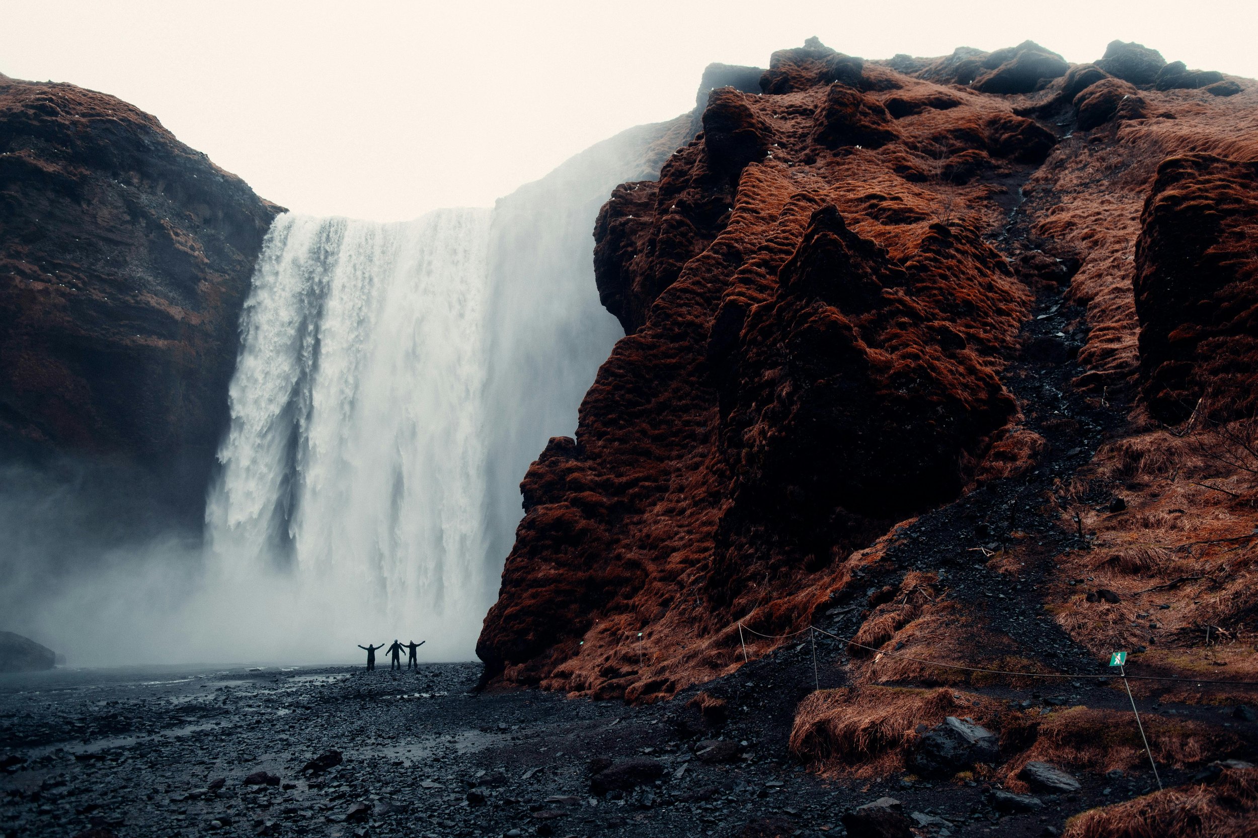Two people standing with arms outstretched near a large waterfall surrounded by rocky, reddish-brown terrain