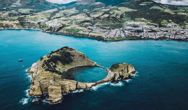 A volcanic crater with surrounding rocky cliffs situated in the ocean near a coastal town, with hills and farmland in the background.