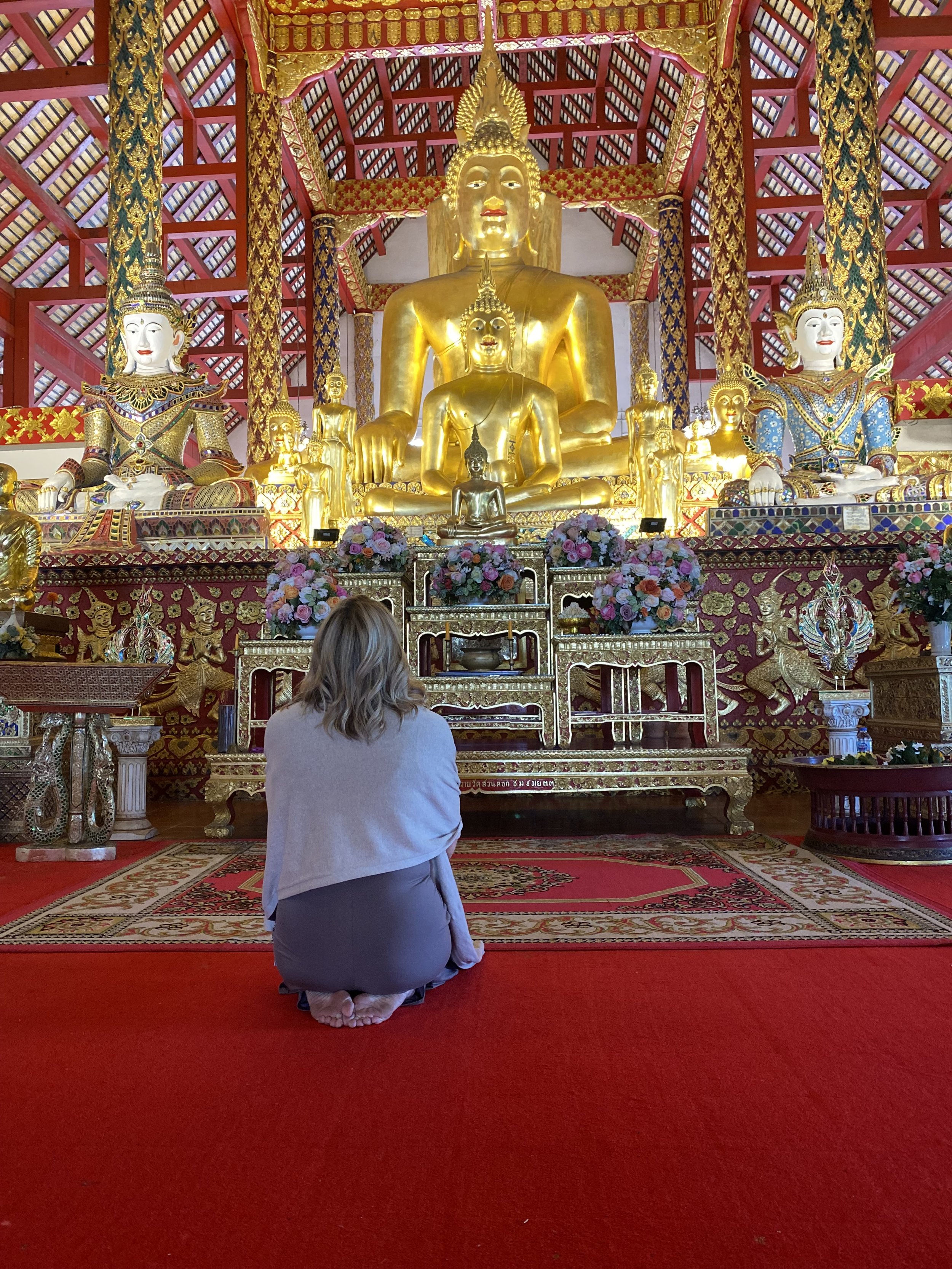 A woman kneeling in prayer before a large golden Buddha statue and smaller statues inside a richly decorated temple with red carpet, floral arrangements, and intricate patterns.