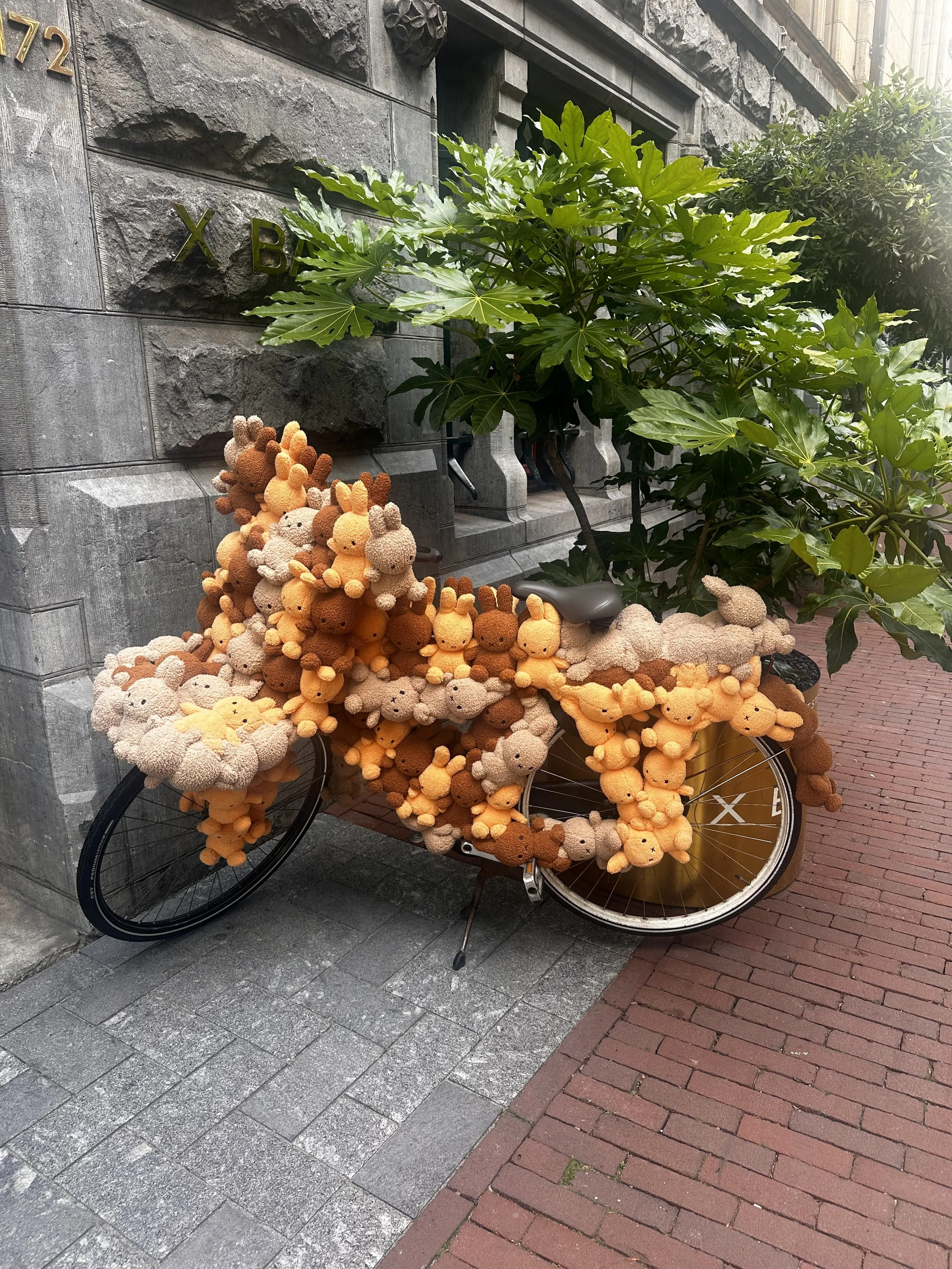 Bicycle decorated with numerous plush stuffed animals resembling small bears in shades of beige, orange, and brown, parked against a gray stone building with ivy and greenery nearby.