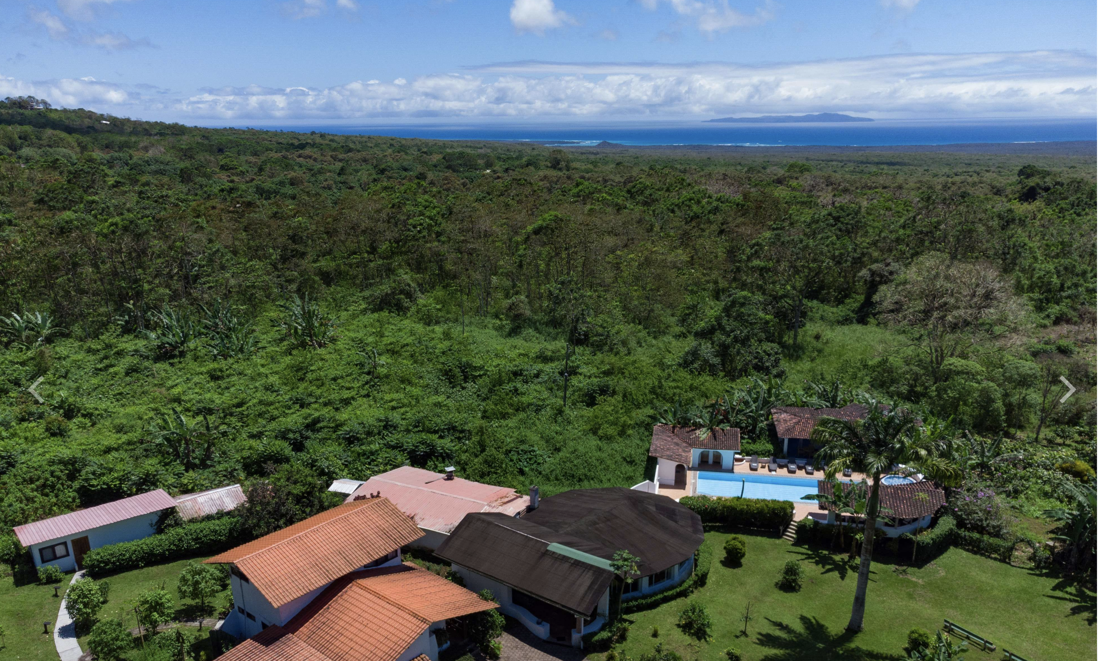 A view of a coastal landscape with lush green trees and residential houses with red-tiled roofs in the foreground, a dense forest extending to the horizon, and the ocean with an island in the distance under a partly cloudy sky.