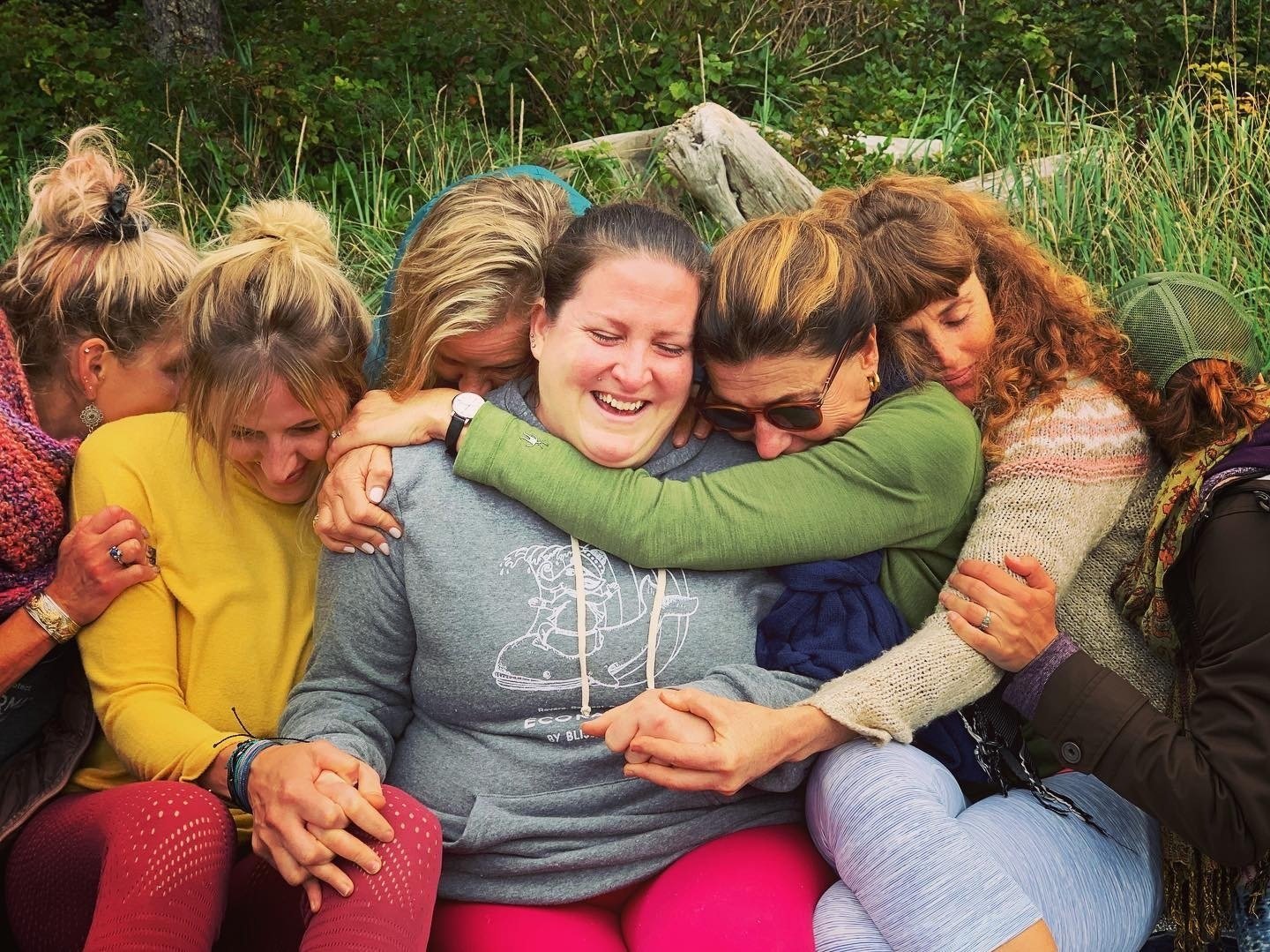 Group of women hugging and laughing outdoors surrounded by greenery.