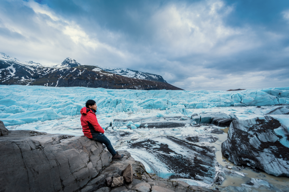 A person in a red jacket sitting on a rock near a glacier with snow-capped mountains in the background under cloudy skies.