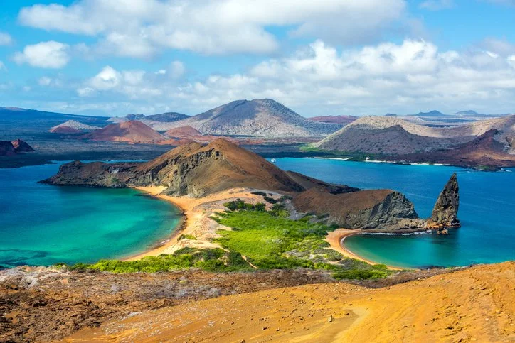 A view of a volcanic caldera with a blue lake, surrounded by colorful rocky terrain and distant mountains under partly cloudy skies.