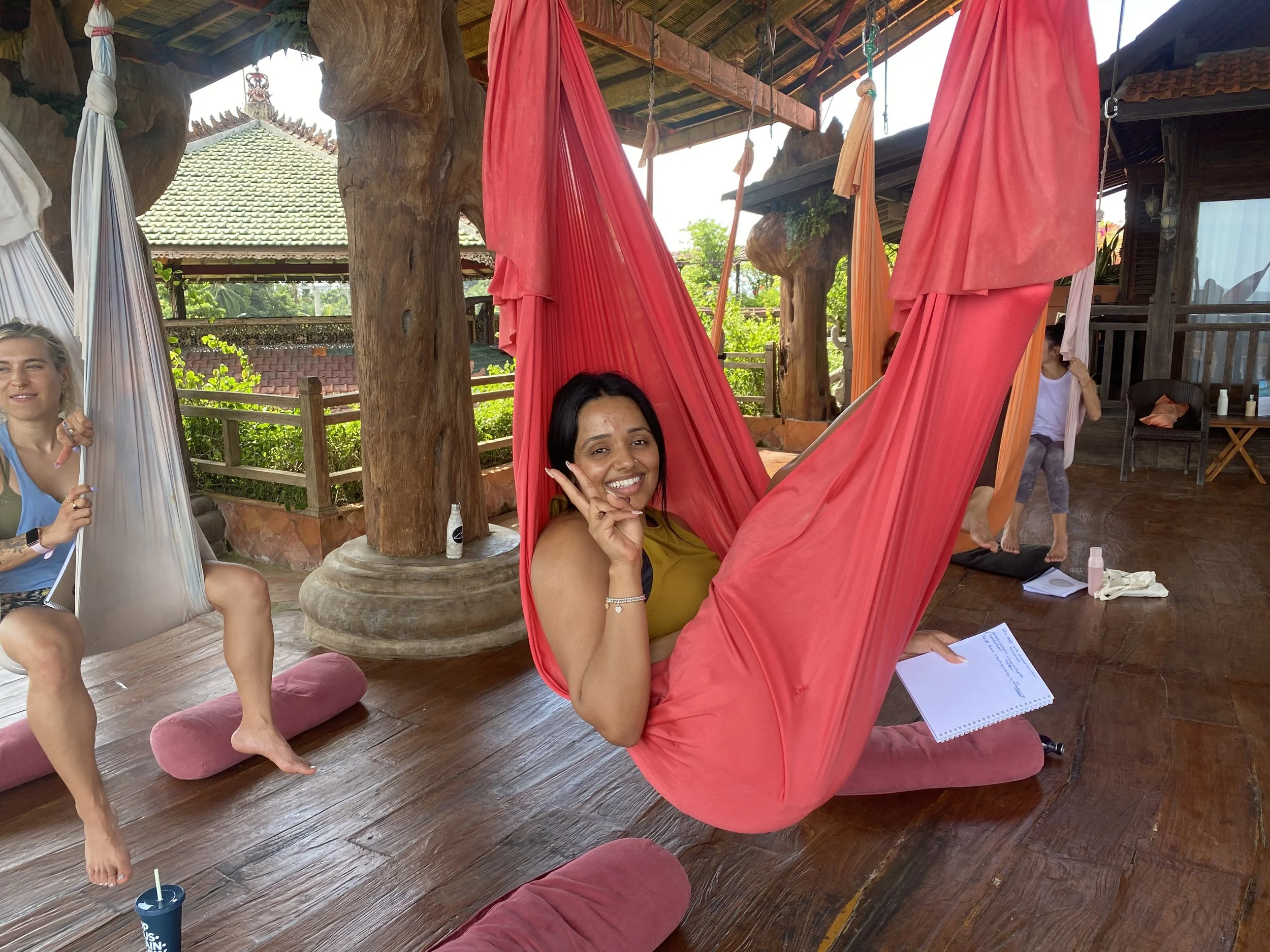 A group of women practicing yoga outdoors on wooden floor under a covered structure with large wooden supports, surrounded by greenery. One woman in the foreground is smiling and making a peace sign while sitting in a hammock-like yoga swing. Other women are sitting or standing in background, with yoga mats and water bottles nearby.