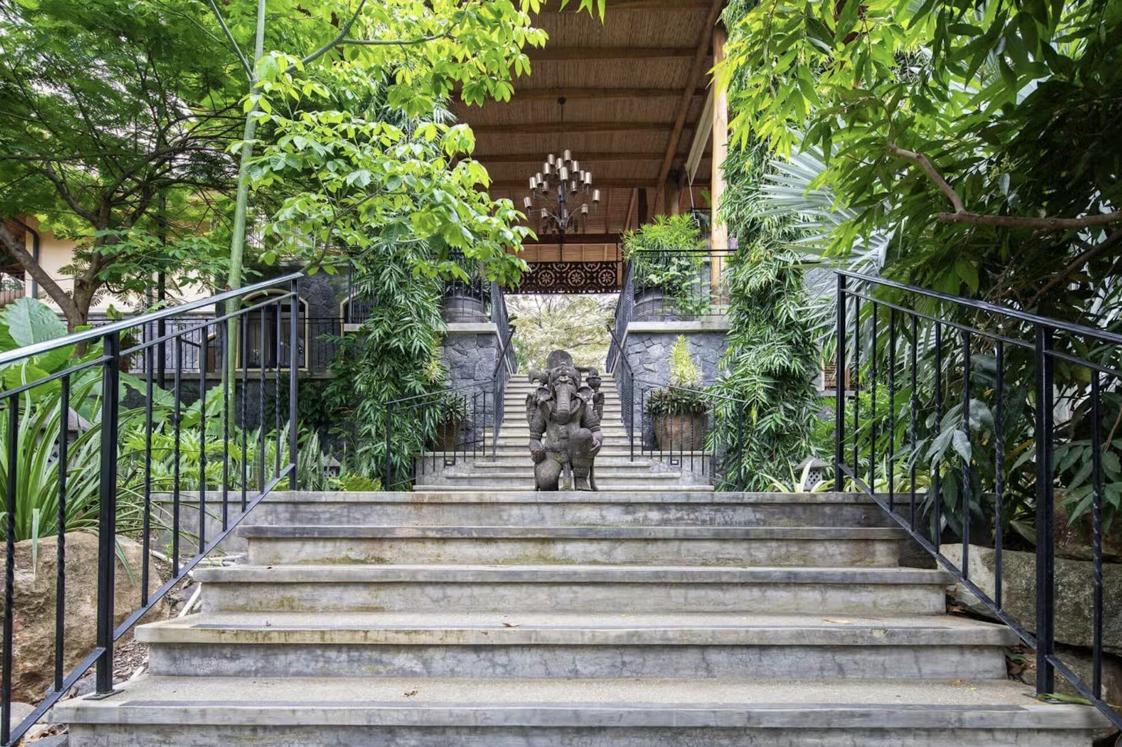 Stone staircase surrounded by lush green plants leading to a porch with a wooden roof, a chandelier, and a statue of multiple elephants at the top of the stairs.