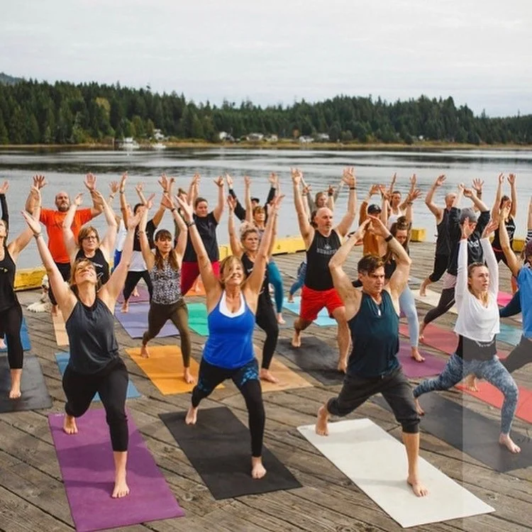 People practicing yoga outdoors on a dock near a lake, with trees and a cloudy sky in the background.