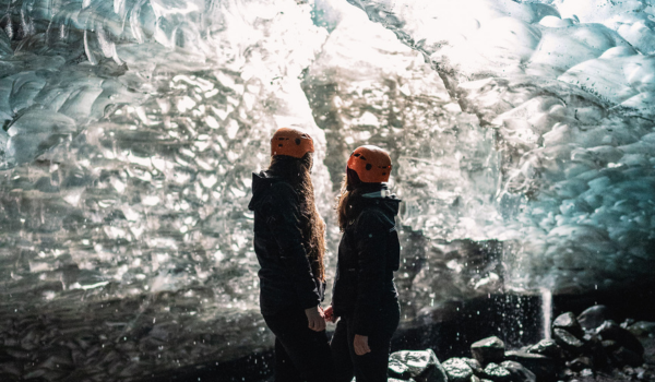 Two people in helmets standing inside an ice cave with ice formations overhead.