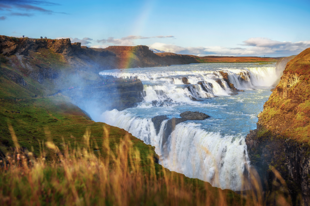 Large waterfall cascading over cliffs with a rainbow in the mist and a blue sky with some clouds