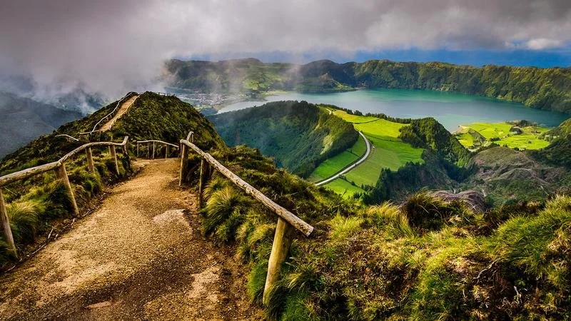 A dirt hiking trail with wooden railings winding along a lush green mountainside overlooking a lake and valley.