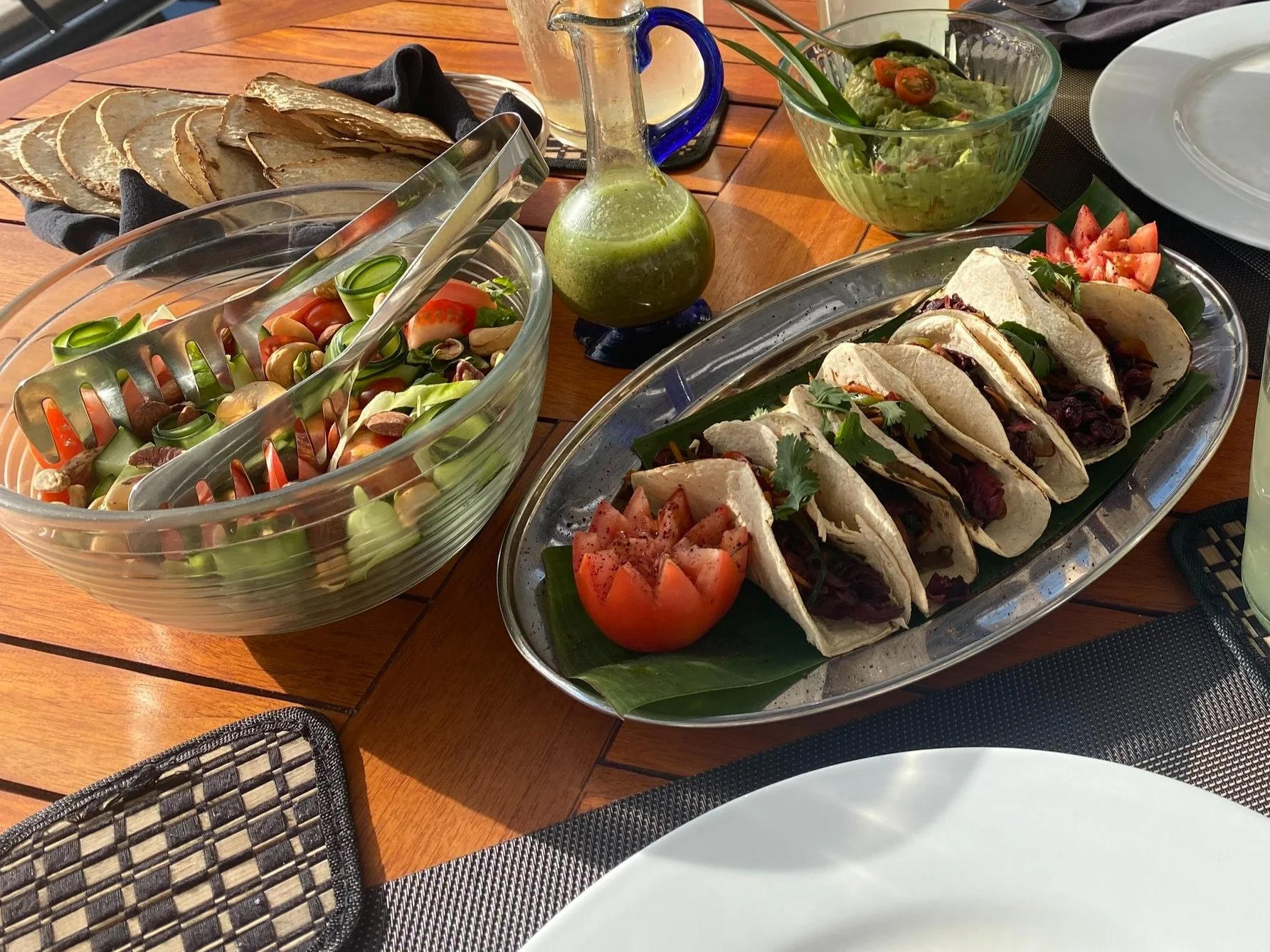 A table with a large bowl of vegetable salad, a platter of tacos, a bowl of guacamole, and a plate of sliced tomatoes.
