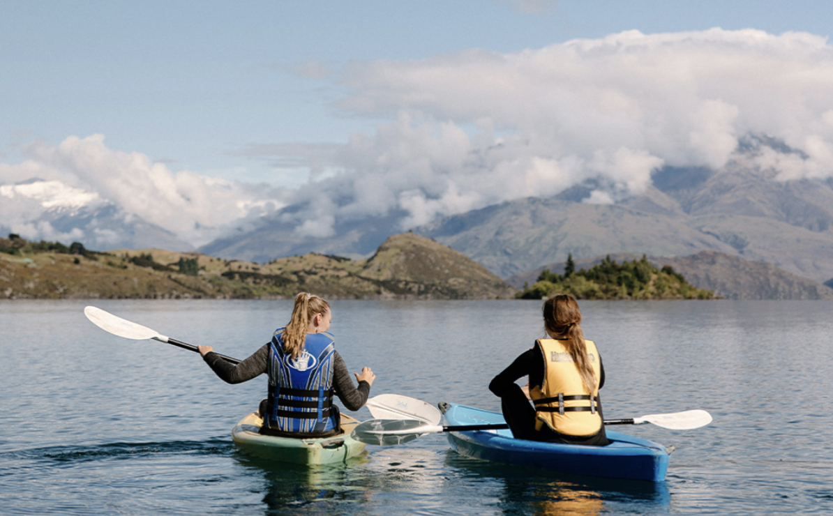 Two women kayaking on a calm lake with mountains and cloudy sky in the background.