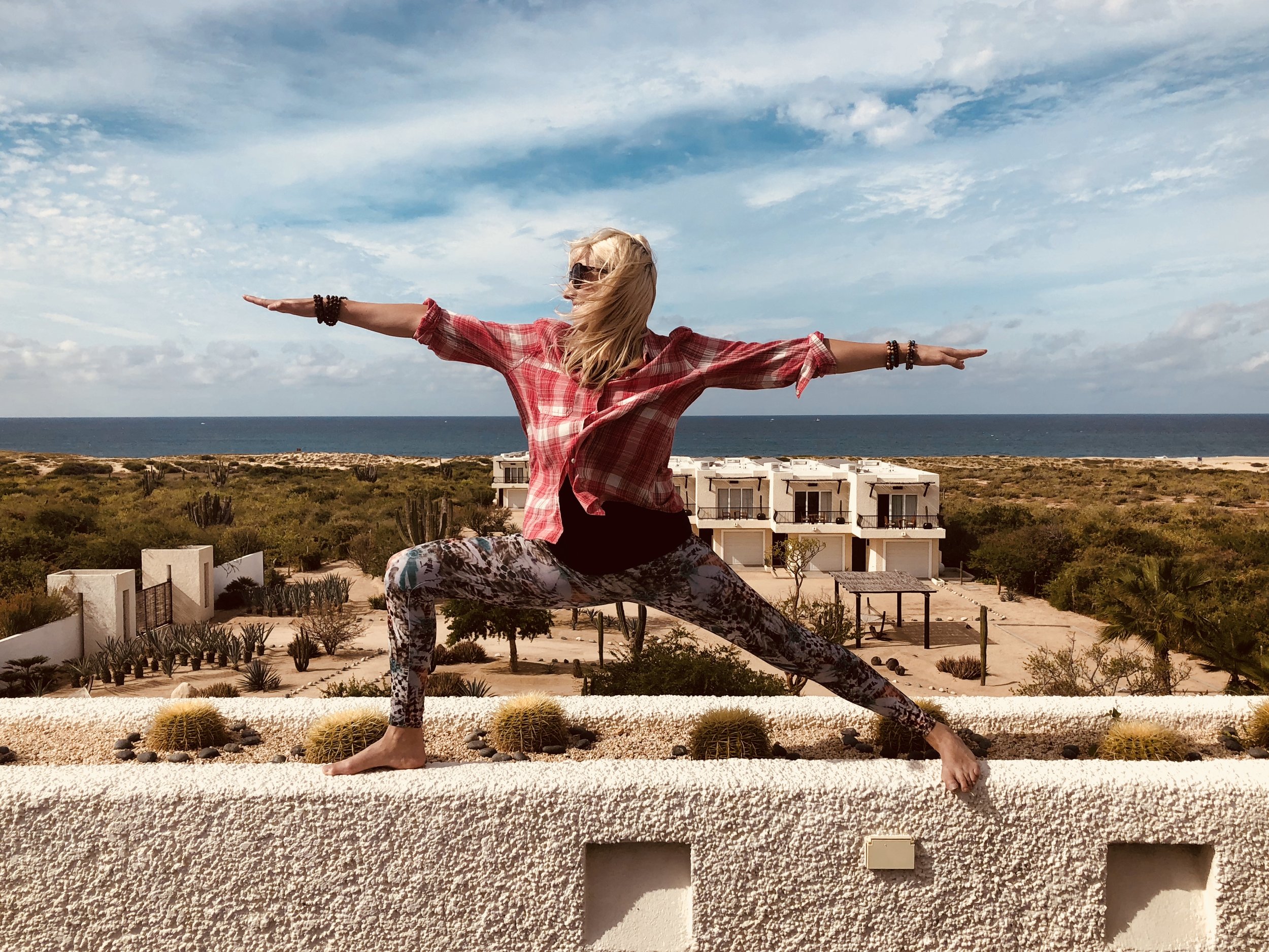 A woman practicing yoga on a rooftop ledge with a desert landscape and ocean in the background.