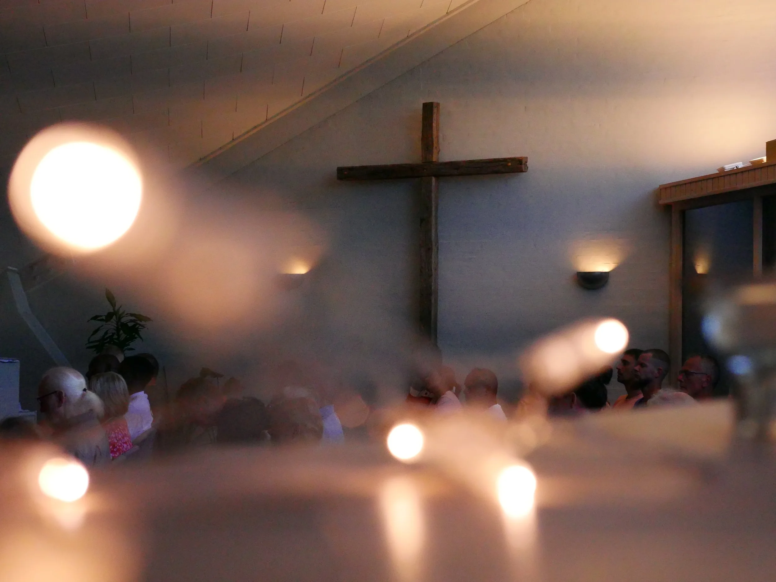 Billedet viser en forsamling mennesker i en kirke, med en stor simpel trækors på væggen i midten. Der er dæmpet belysning, og nogle små lys kan ses i forgrunden.