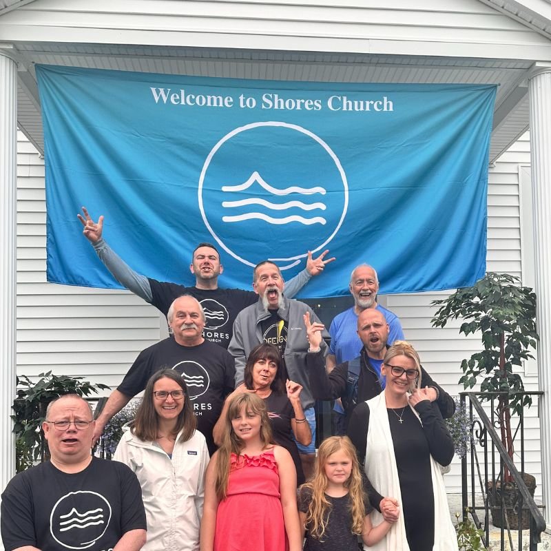 A group of people standing in front of a blue banner that reads "Welcome to Shores Church" and features a wave logo. The group includes adults and children, with some making cheerful gestures and smiling.