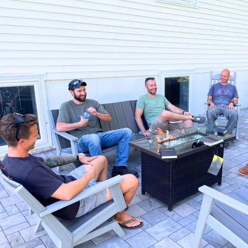 Four men sitting around a fire pit, smiling and talking, on a patio outside a house with white siding.