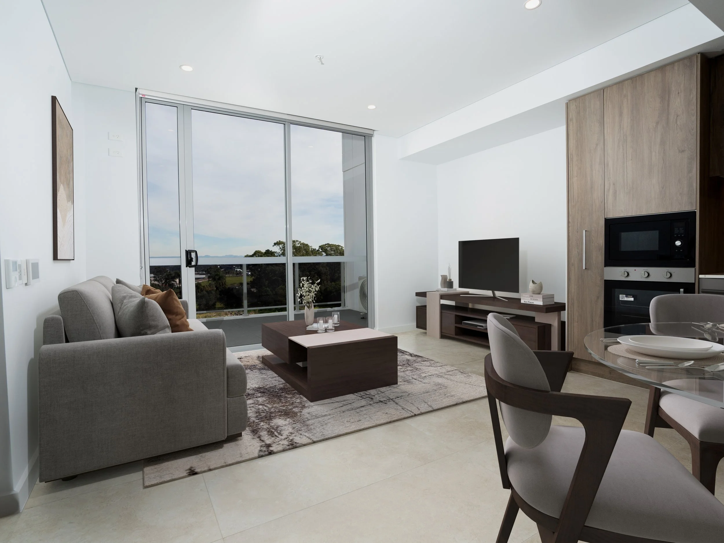 Modern living room with gray sofa, glass sliding door to balcony, TV on wooden stand, dining table with chairs, and a view of trees and sky outside.