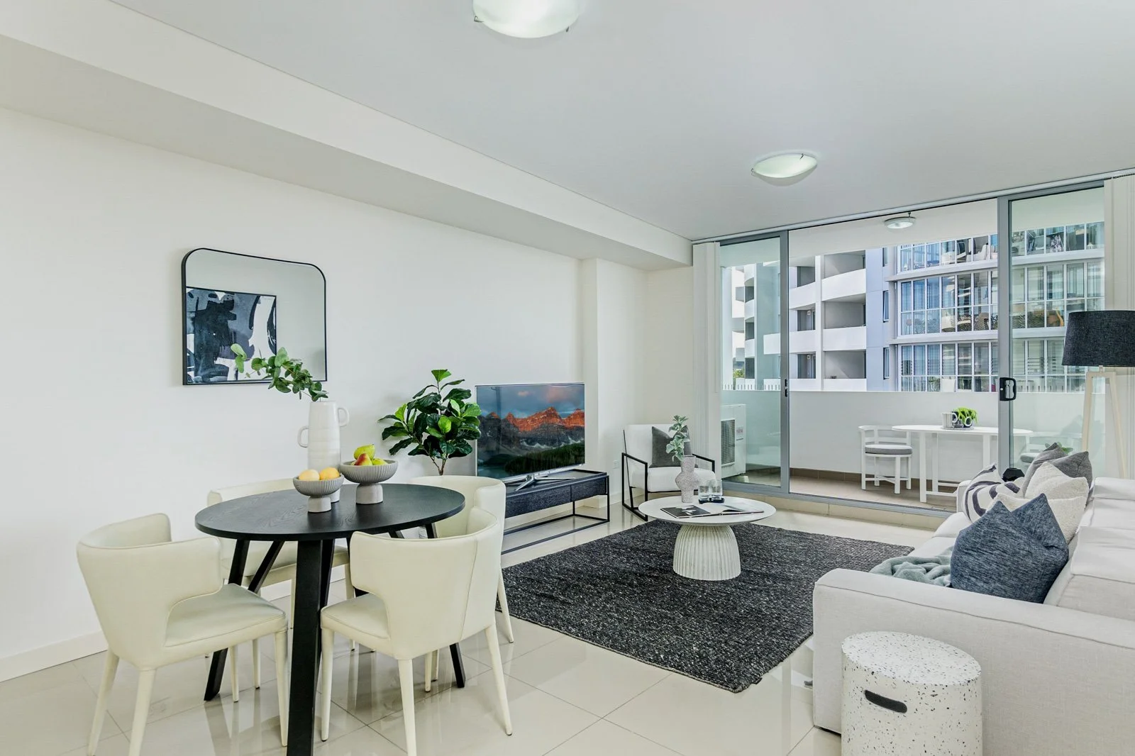 Modern living room with white walls, black and white furniture, a TV, indoor plants, a black rug, and a glass sliding door leading to a balcony with city view.