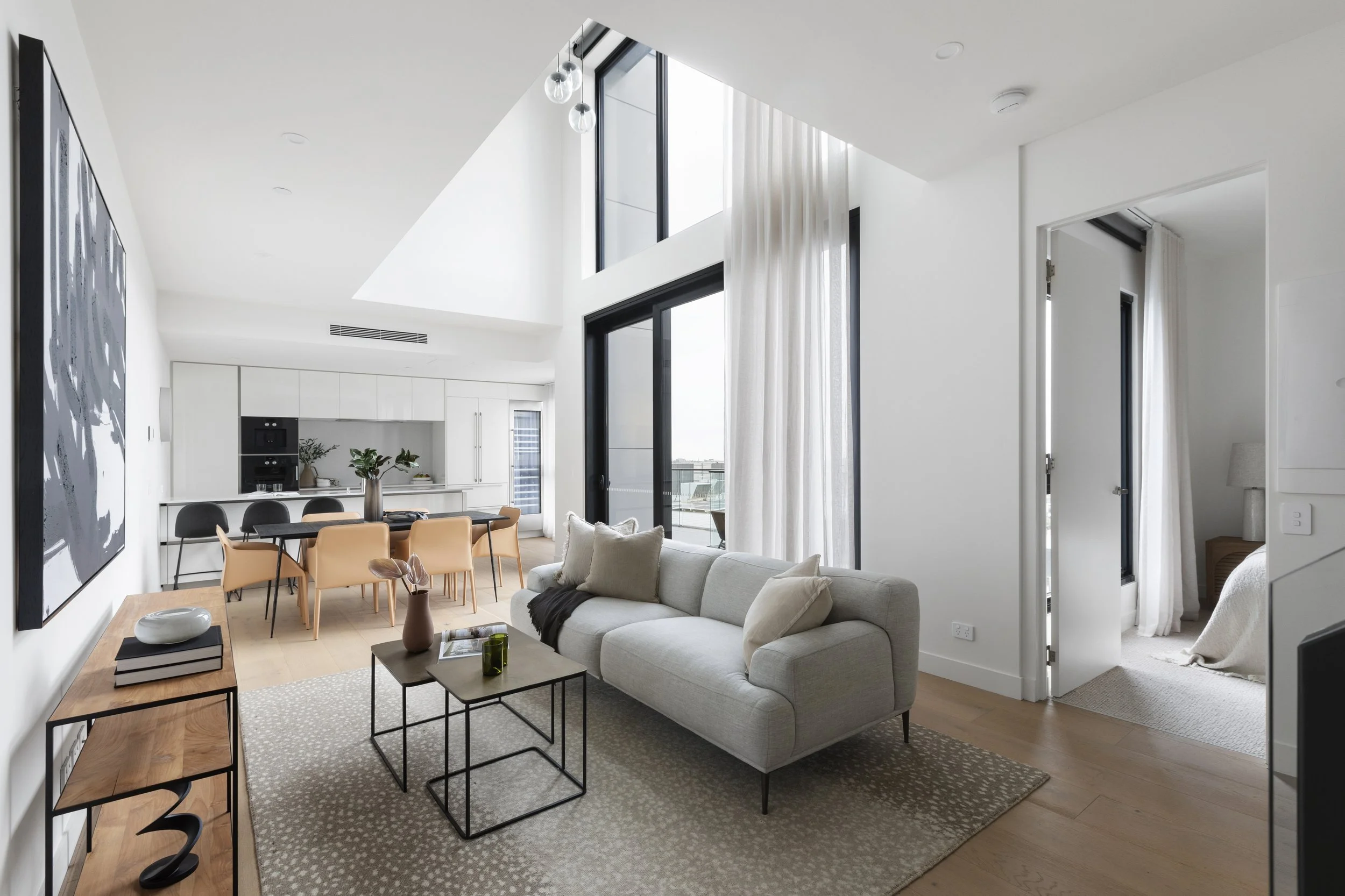 Modern open-concept living and dining area with white walls, large windows, a white sofa, a wooden console table, and a view into a bedroom.