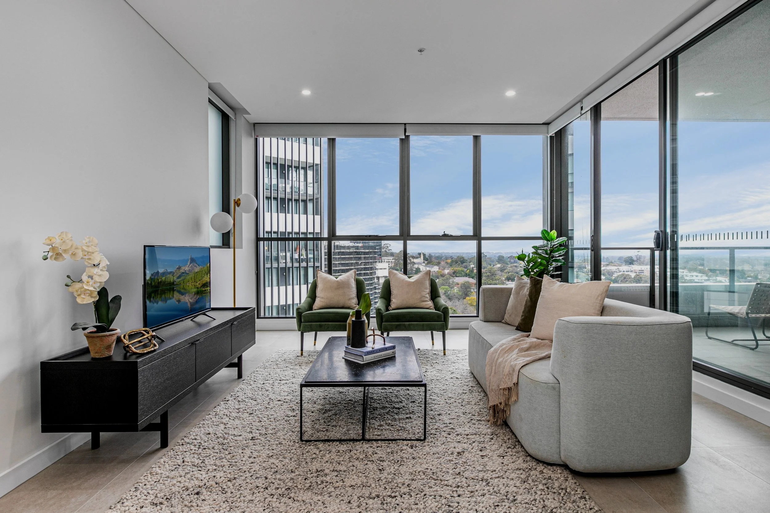 Modern apartment living room with large windows, gray sofa with pillows, green armchairs, black coffee table, TV on black stand, and balcony view of cityscape and sky.