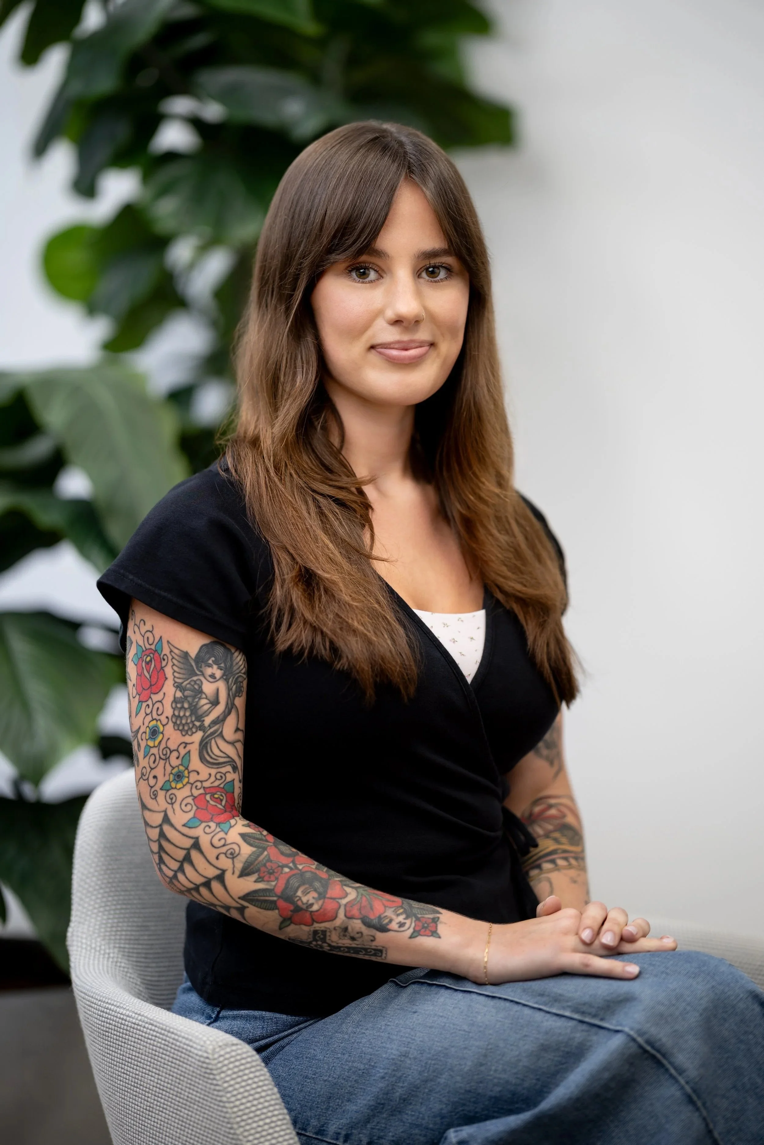 A woman with long brown hair, wearing a plaid dress with short ruffled sleeves, sits on a beige couch in a room with large windows and green plants in the background.