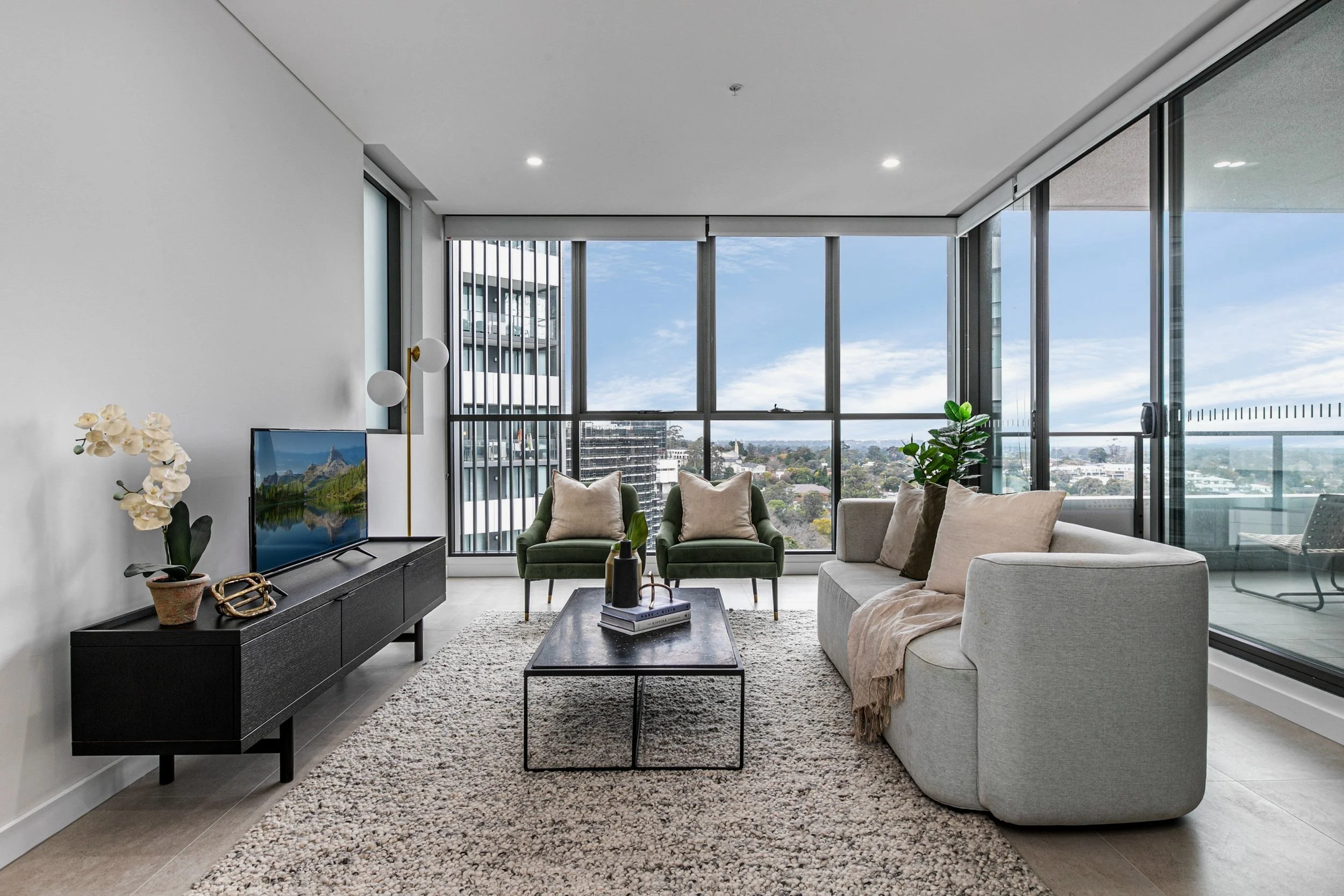 Modern living room with large windows, beige and green seating, black coffee table, TV stand, and balcony view.