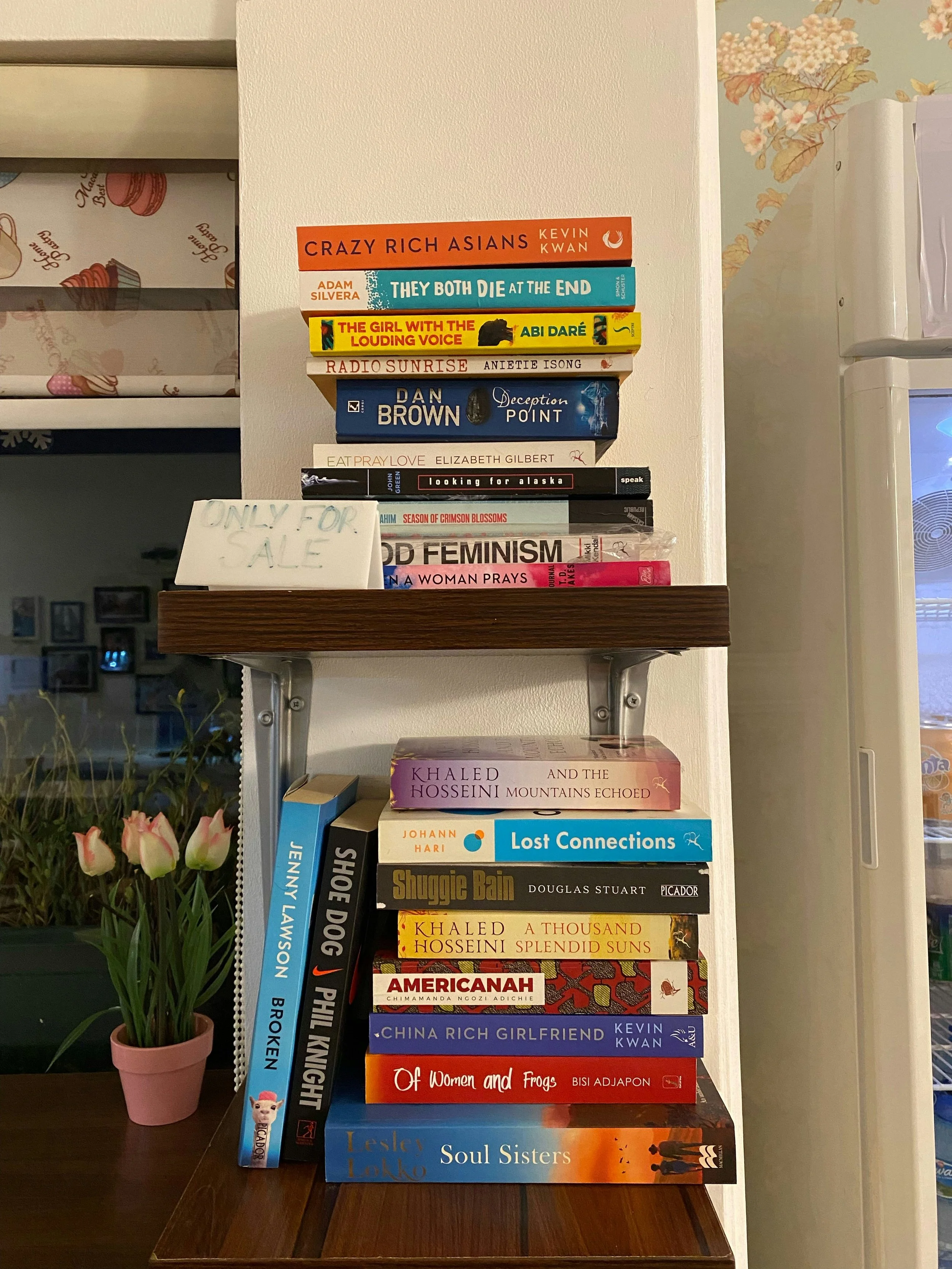 Stack of books on a wooden table and a shelf, with more books stacked on top. The books include titles such as 'Crazy Rich Asians', 'They Both Die at the End', 'The Girl with the Louding Voice', 'Radio Sunrise', 'Deception Point', 'Eat Pray Love', 'Looking for Alaska', 'God Feminism', 'A Woman Prays', 'And the Mountains Echoed', 'Lost Connections', 'Shuggie Bain', 'A Thousand Splendid Suns', 'Americanah', 'China Rich Girlfriend', 'Of Women and Frogs', and 'Soul Sisters'. There's a small potted plant with pink flowers on the table, and a white sign that reads 'Only for Sale'.