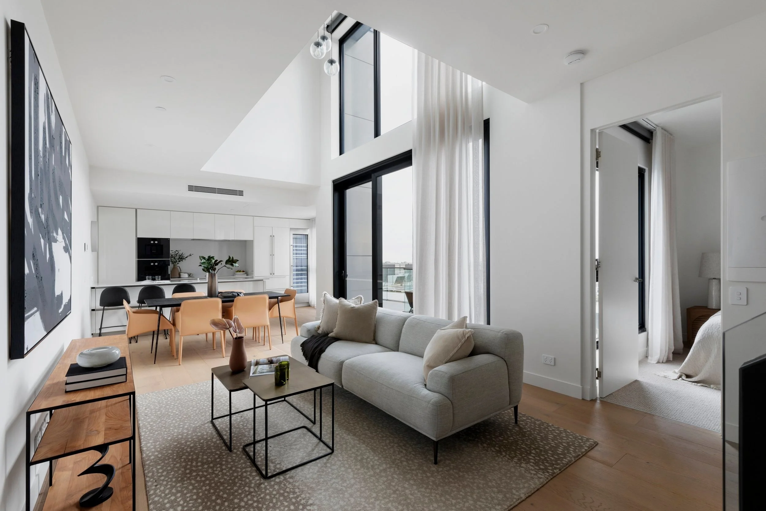 Living room with beige sofa, coffee tables, and patterned rug, open kitchen and dining area with black and tan chairs, large windows with white curtains, and a bedroom visible through an open door.