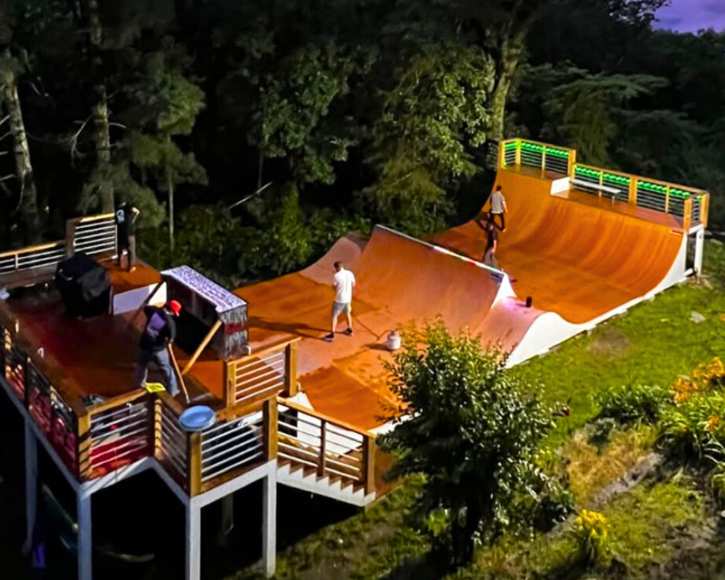 People constructing or maintaining a wooden skatepark with ramps and rails at night, surrounded by trees and greenery.