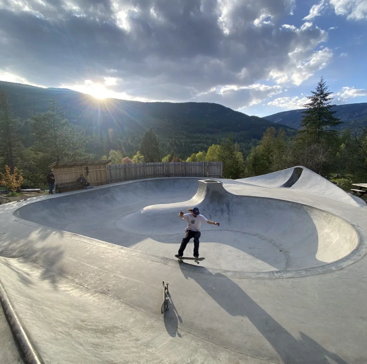 Skateboarder in a concrete skate park with mountains and trees in the background, during sunset.