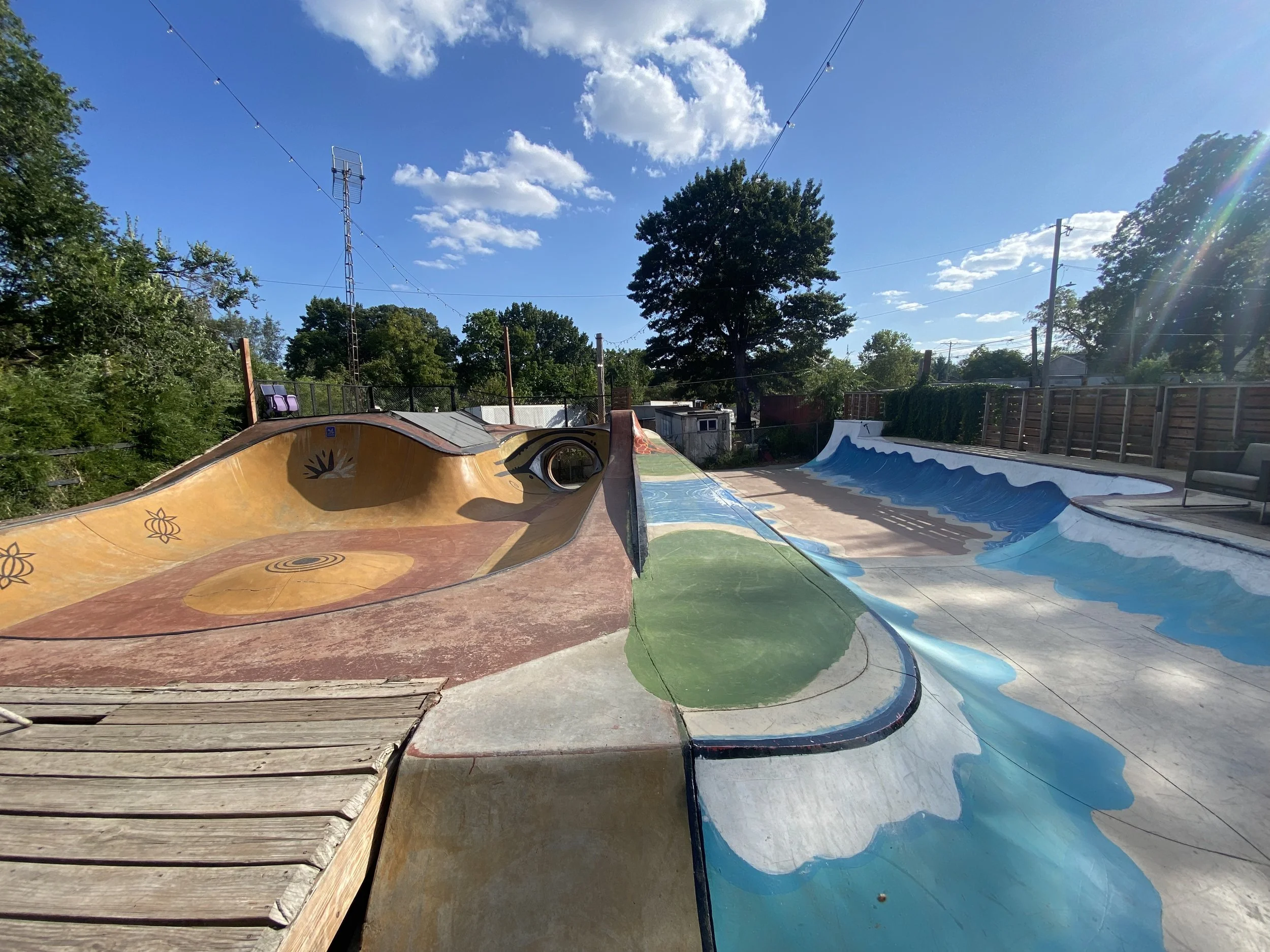 Empty outdoor skatepark with colorful painted ramps and bowls under a blue sky with clouds, surrounded by trees and a wooden fence.
