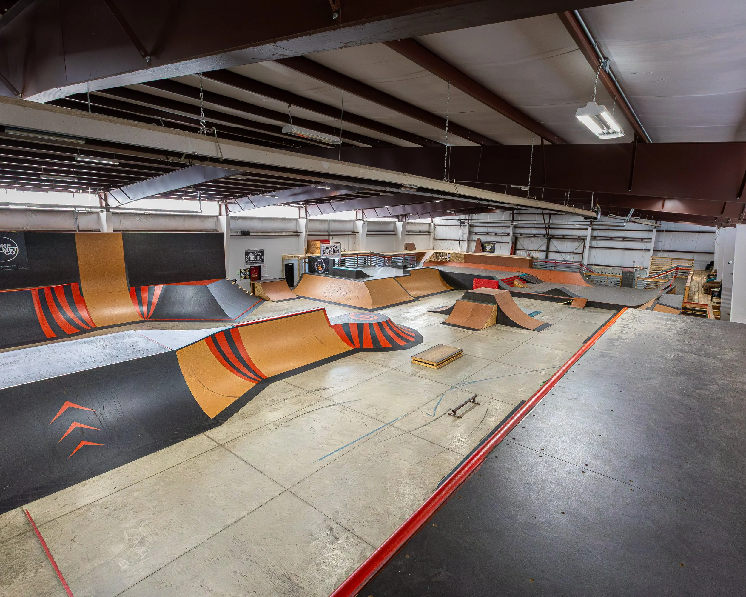 Indoor skatepark with ramps, rails, and obstacles for skateboarding and BMX riding, featuring a concrete floor and metal roof.