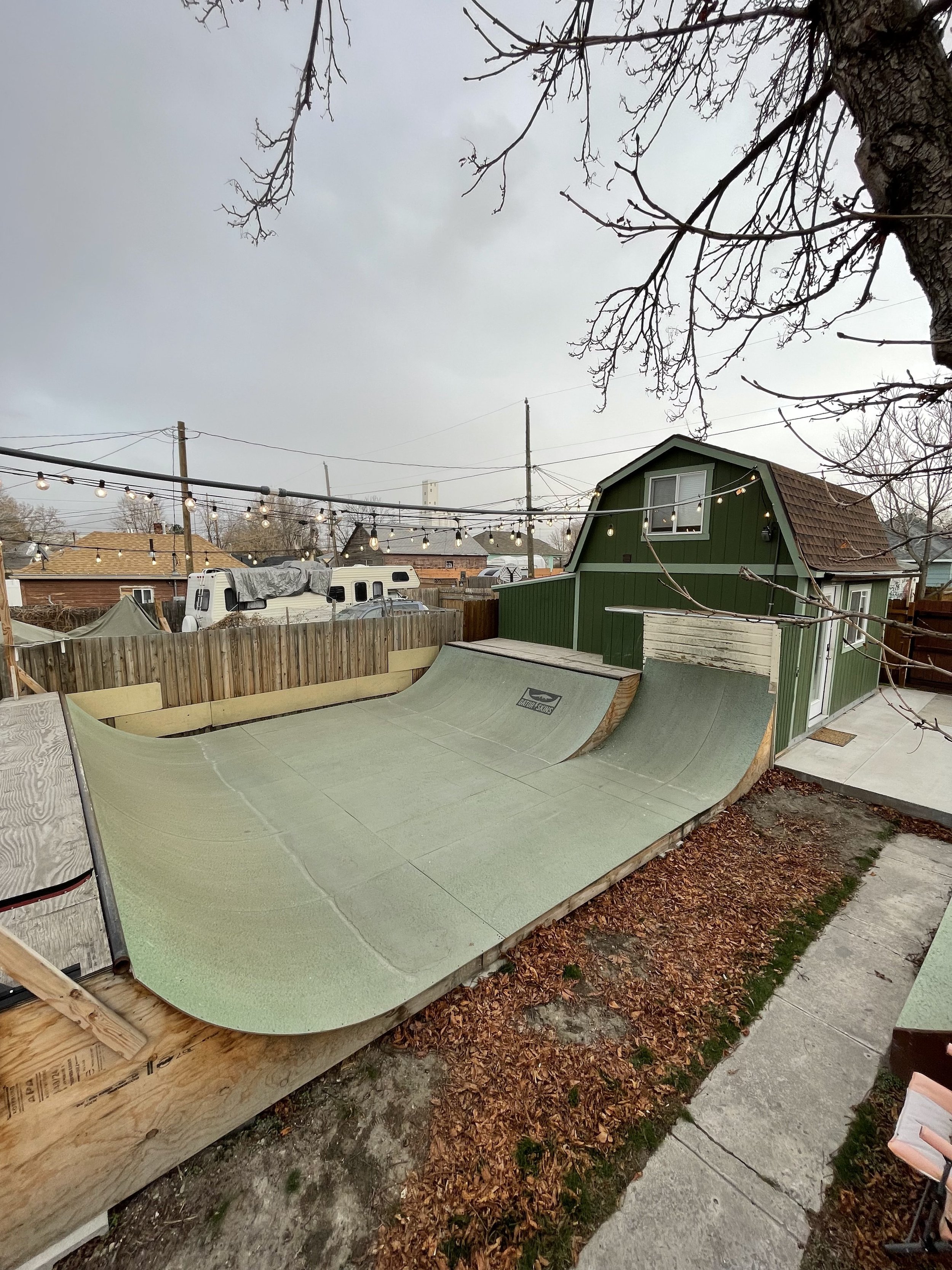 Backyard with a miniature skate ramp, a green house with an upper window, string lights, leafless tree, and RVs in the background.