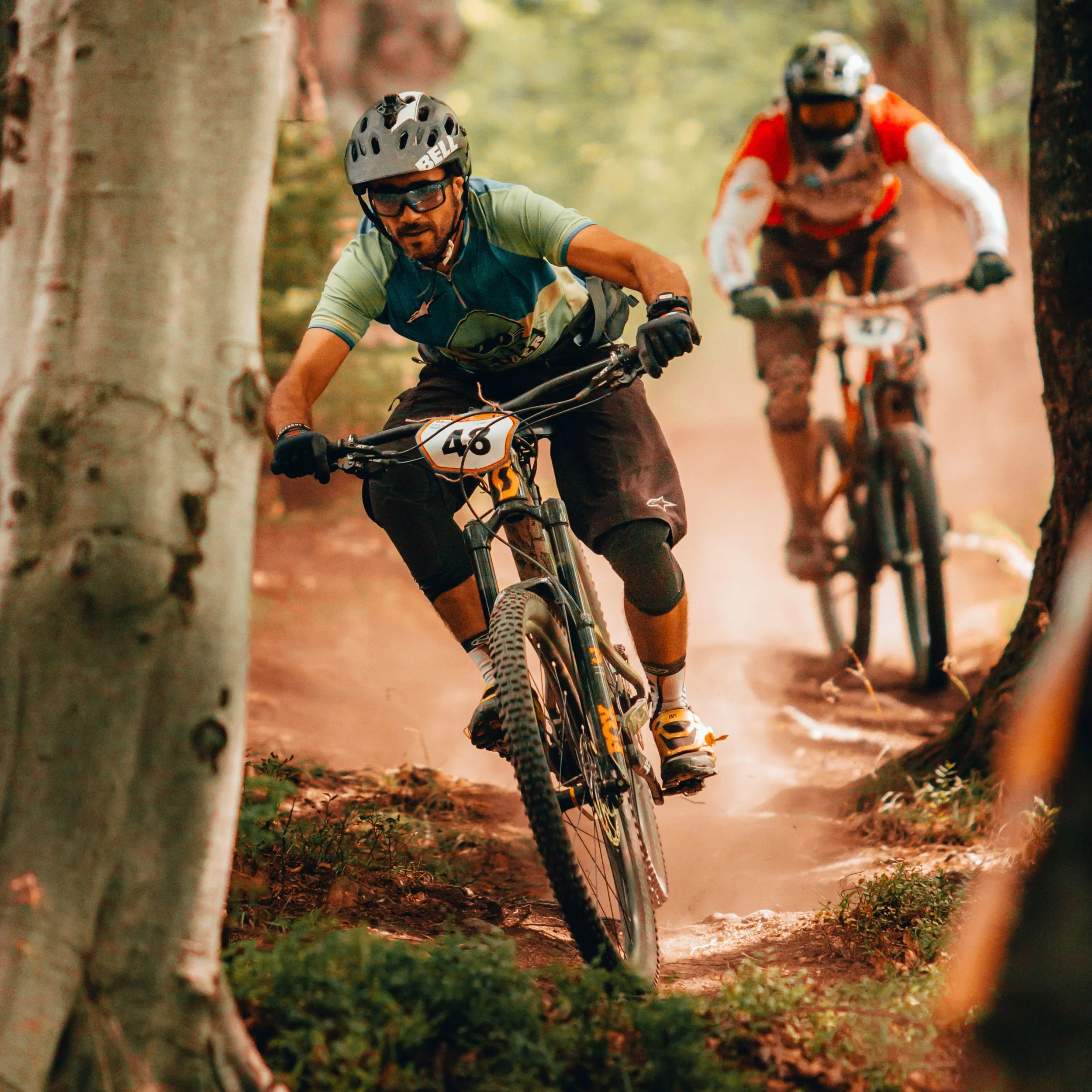 Two mountain bikers racing on a dirt trail through a forest with trees on both sides, kicking up dust.