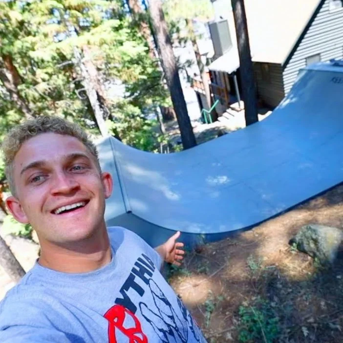Garrett Ginner smiling and taking a selfie outdoors near a skate ramp, with trees and a house in the background.