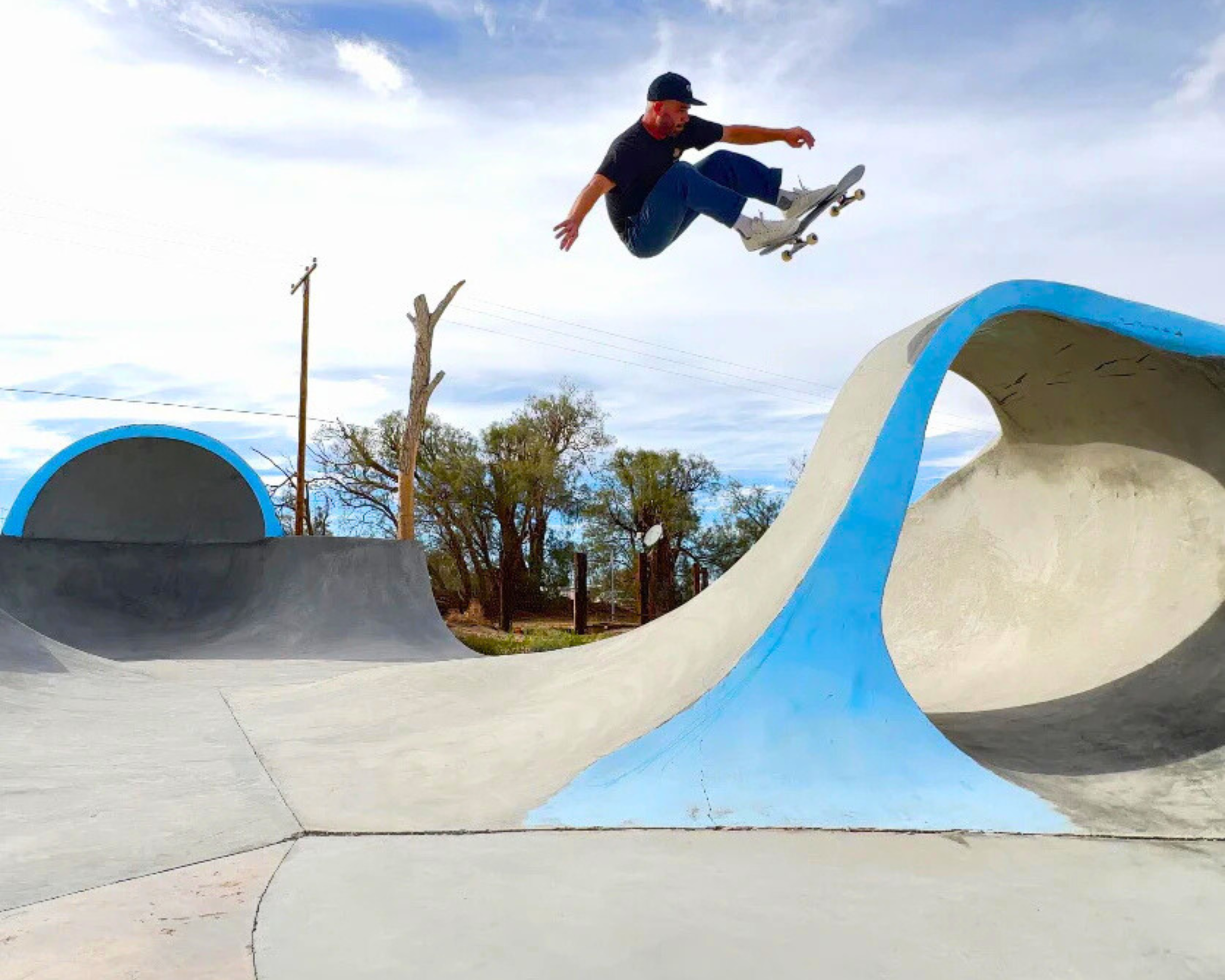A skateboarder is airborne at a skate park with curved ramps painted in blue and white, trees, and a partly cloudy sky in the background.