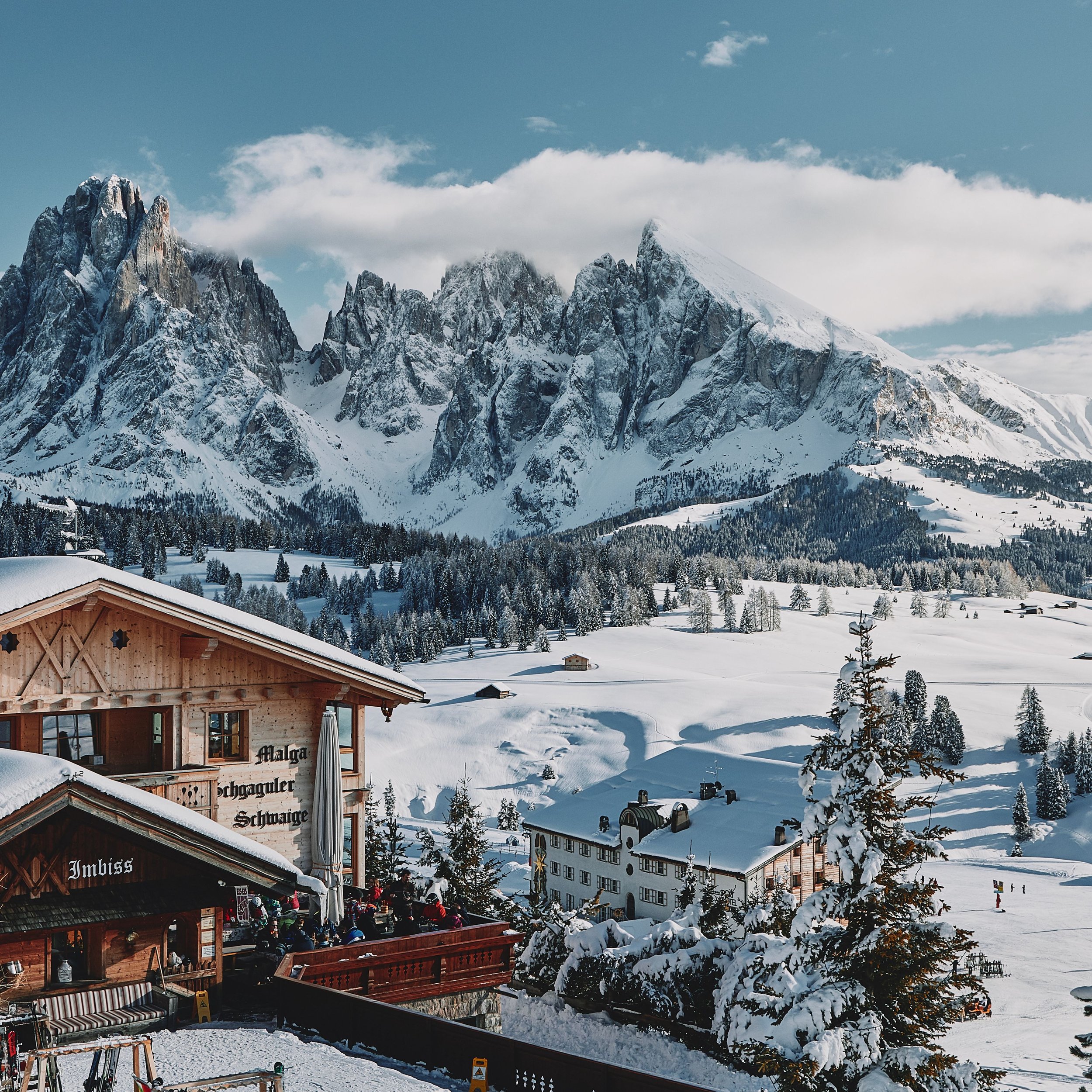 Snow-covered alpine village with chalet-style buildings, surrounded by snowy trees and mountains under a partly cloudy sky