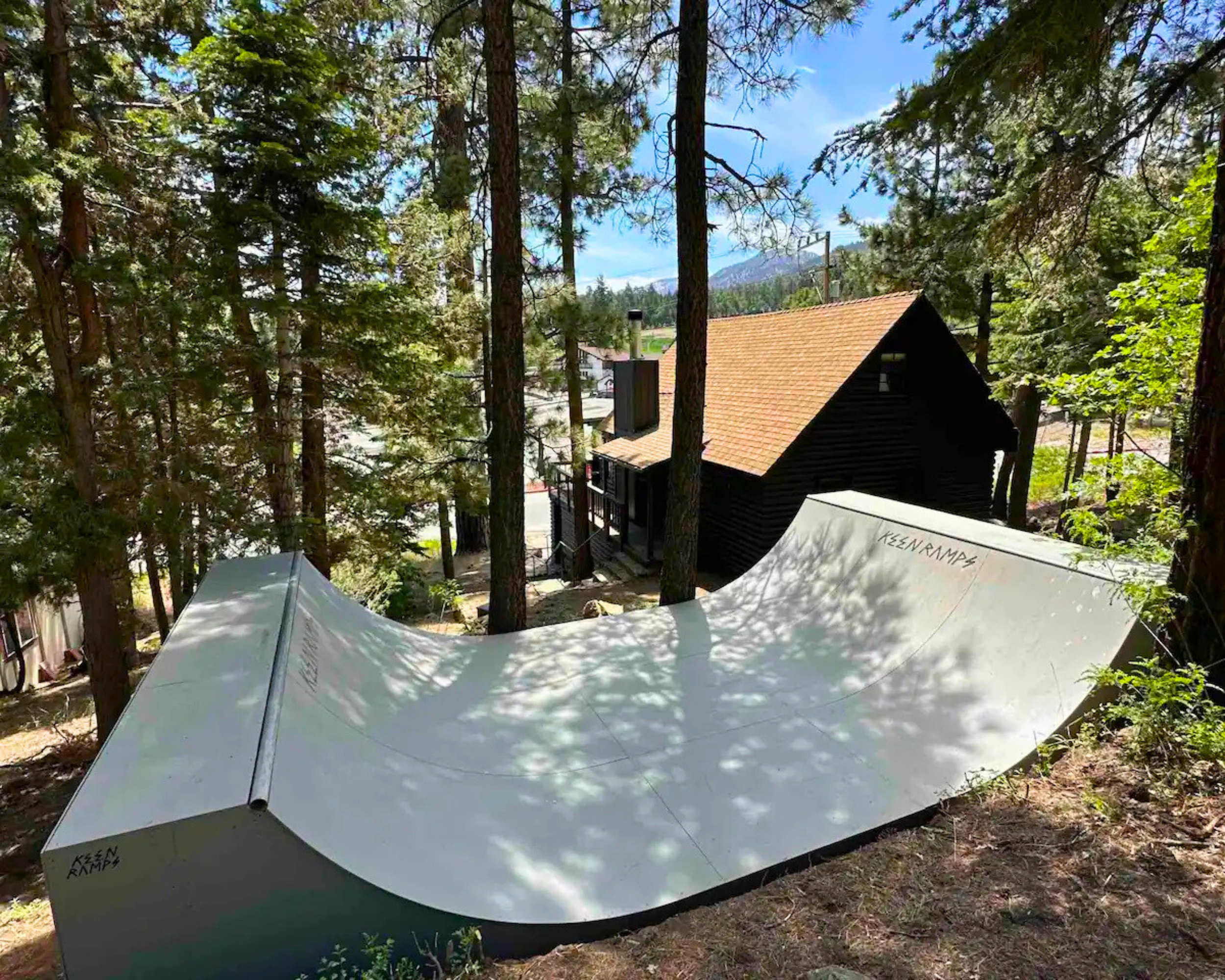 A white skate ramp set in a wooded area with tall trees, with a dark wooden house and mountains visible in the background.